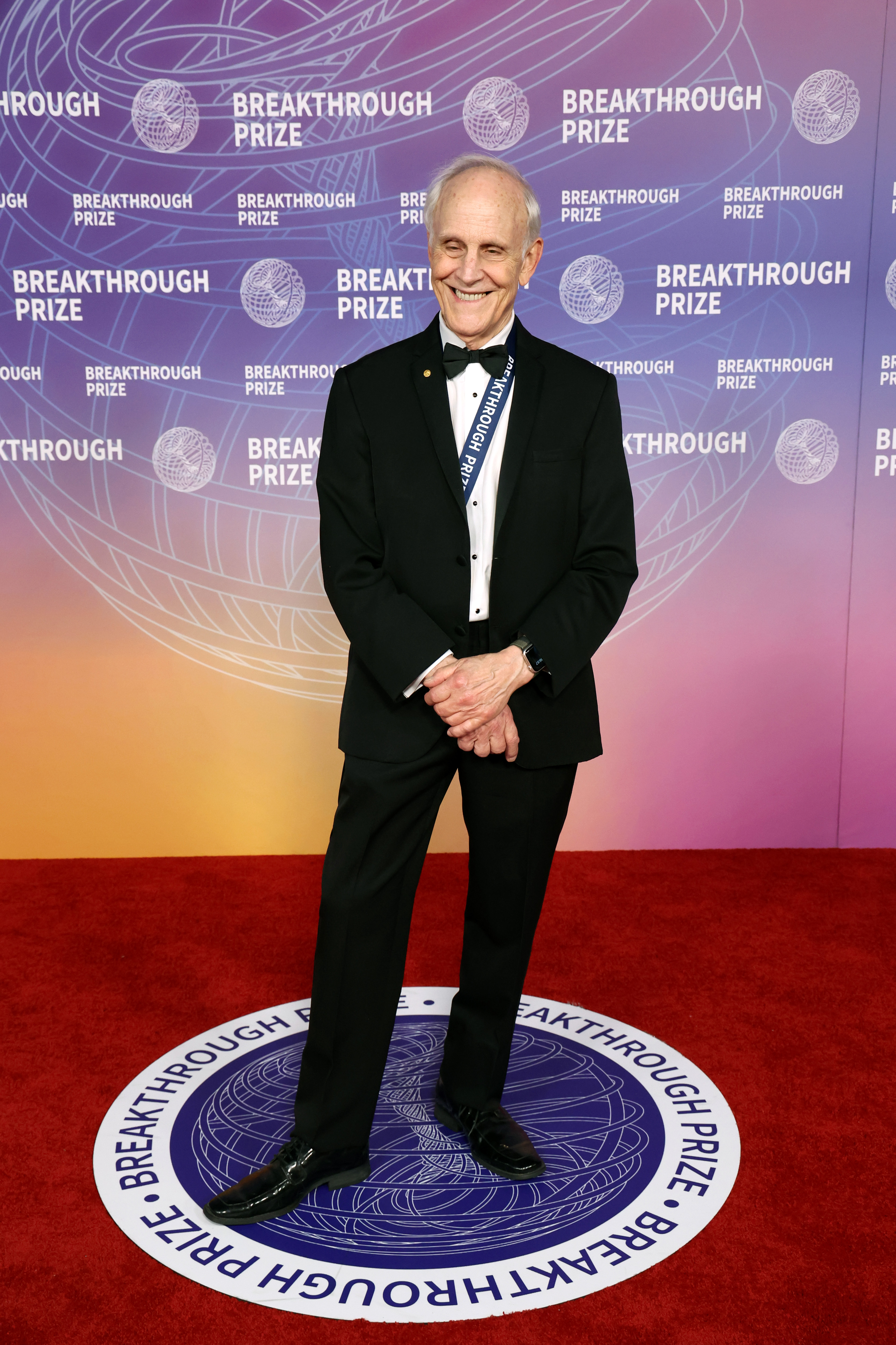 David Gross in a black tuxedo and “BREAKTHROUGH PRIZE” sash standing on a red carpet before a purple Break Through Prize event backdrop.