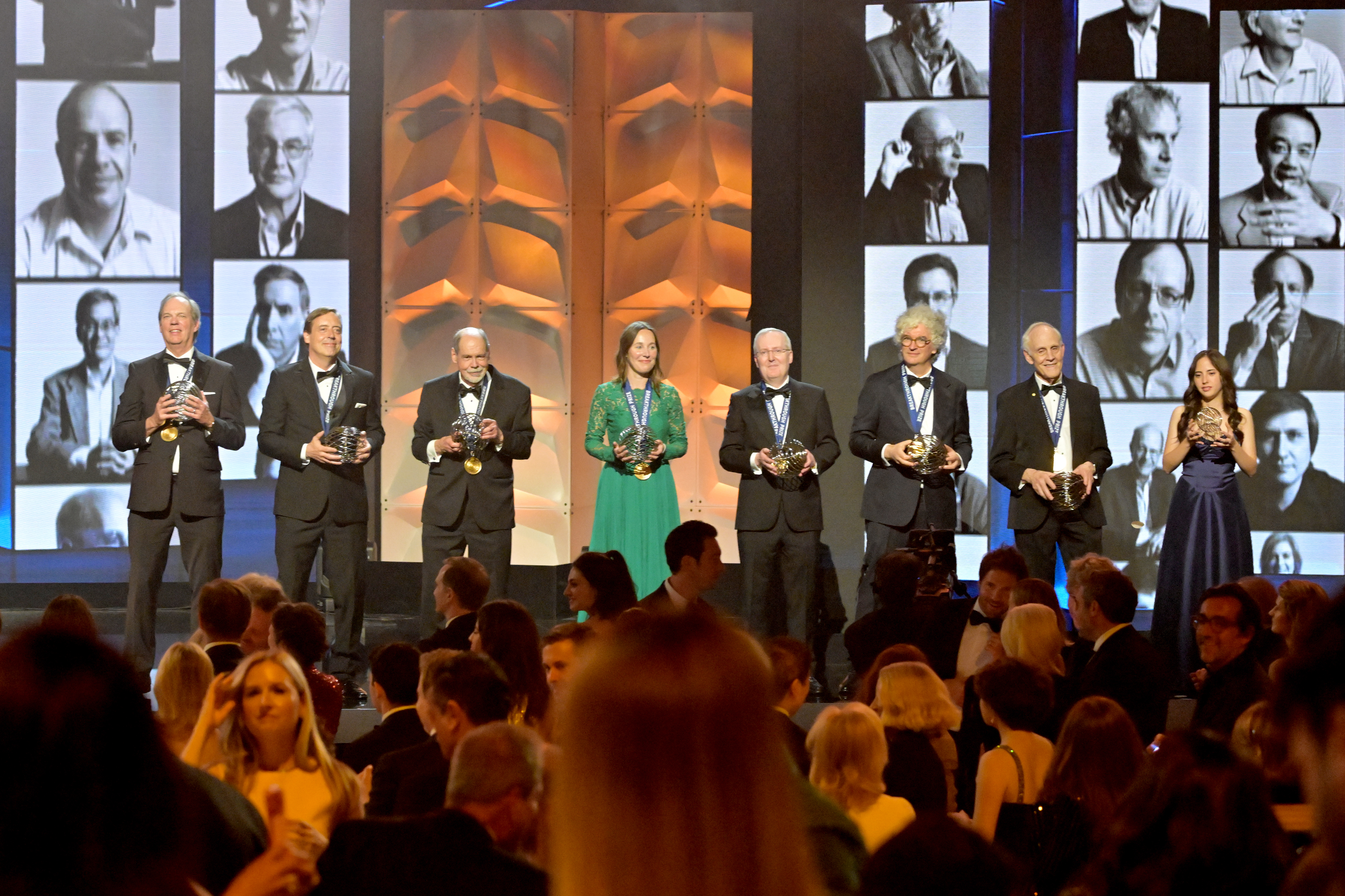 Eight award recipients, including David Gross, pose onstage with trophies as a seated audience watches, surrounded by orange geometric panels and large black-and-white portrait displays.