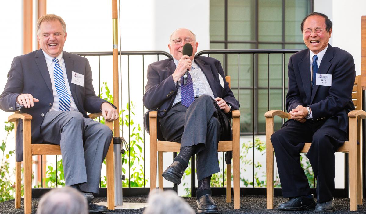 three men seated on a stage, Charlie in the center is holding a microphone